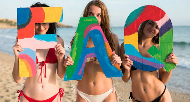Trois étudiantes sur la plage tiennent des lettres FAQ colorées lors d’un séjour linguistique LISA! à Malte