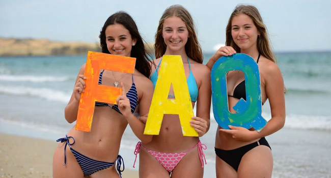  Jeunes adultes sur la plage à Majorque tiennent des lettres FAQ lors d’un séjour linguistique LISA! avec la mer en arrière-plan