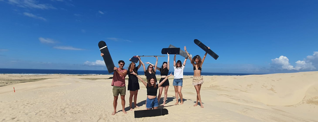 Mehrere Menschen posieren mit erhobenen Händen und Skateboards am Strand vor dem Meer in Sydney