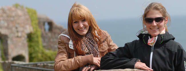 Ausflug ans Meer bei einer LISA! Sprachreise nach Jersey, England Zwei Frauen lächseln vor dem Meer und alten bewachsenen Steinen in die Kamera bei einer Sprachreise Jersey