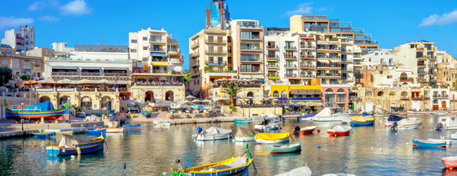 Blick auf den Hafen von St. Julian's, Malta, mit bunten Fischerbooten und modernen Gebäuden im Hintergrund.