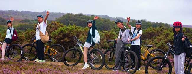 Schüler auf Fahrrädern winken in die Kamera vor einer englischen Landschaft